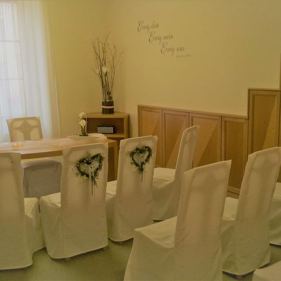 Ein Raum mit einem Tisch und Stühlen, die für einen besonderen Anlass vorbereitet sind. Die Stühle haben weiße Überzüge mit herzförmigen Dekorationen. Es gibt einen hölzernen Schrank mit einer Pflanze oben. Die Wand hat eine dekorative Phrase.