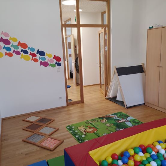 Ein Kinderspielzimmer mit Holzboden, einer bunten Fischwand, einer Spielmatte und einem Ballbecken voller bunter Bälle. In der Ecke steht auch ein Zelt.