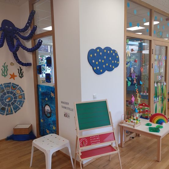 Ein Klassenzimmer mit einem großen blauen Oktopus an der Wand, einem Sternenhimmel und Lernpostern. Es gibt einen kleinen Tisch mit einem Regenbogen, einem Stuhl und einer Tafel. Der Raum hat einen Holzboden und Glastüren.