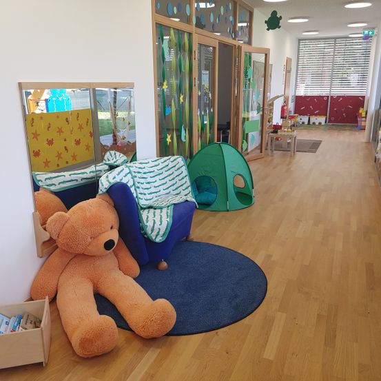 Ein Spielzimmer mit einem Teddybären, einem blauen Teppich, einem blauen Stuhl und einem grünen Zelt. Es gibt auch einen Spiegel, ein Regal mit Büchern und einen Teppich. Die Wände sind weiß mit Dekorationen.