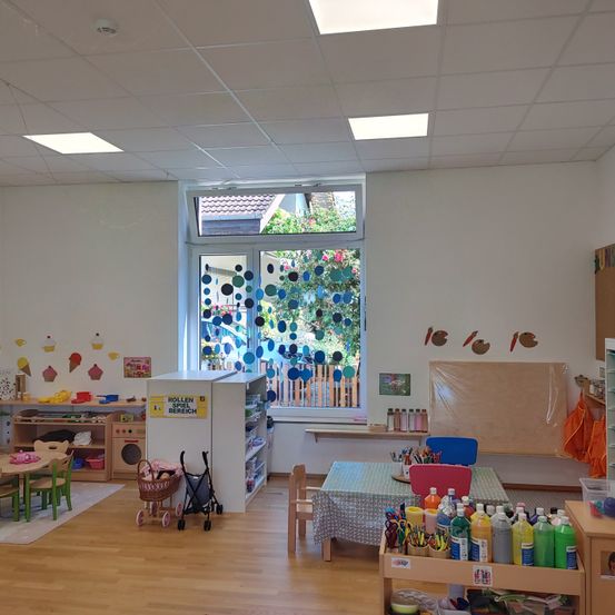 Ein Raum mit Holzboden, weißen Wänden und einem Fenster mit blauen kreisförmigen Dekorationen. Es gibt Tische und Stühle, einen weißen Bücherschrank und Regale mit verschiedenen Gegenständen.