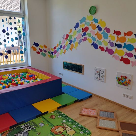 Ein farbenfroher Spielraum mit einer Fischwanddekoration, einem großen Ballbad und verstreuten Spielzeugen auf dem Boden.