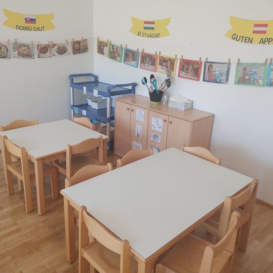 Ein Klassenzimmer mit Holztischen und Stühlen, einem blauen Wagen und einem Holzschrank. Die Wand hat einen gelben Banner mit einer Flagge, eine Reihe von Bildern und einem Regal mit Küchenutensilien.