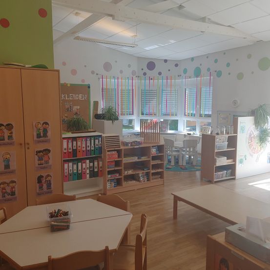 Ein Klassenzimmer mit Holzregalen, bunten Stühlen und einem Tisch mit einem Behälter voller Buntstifte. Die Wände sind mit bunten Punkten bemalt und es gibt Fenster mit Jalousien.