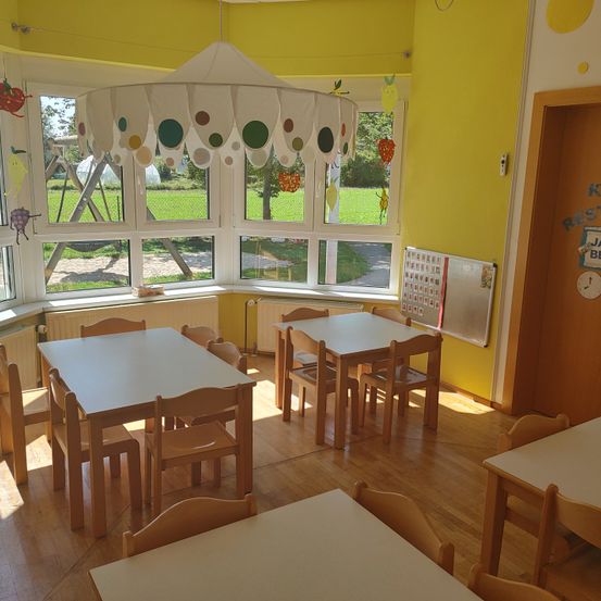 Ein Klassenzimmer mit gelben Wänden, einem runden Fenster und einer hängenden Lampe. Es gibt Tische und Stühle angeordnet und eine Tafel mit Zahlen und Buchstaben. Vor dem Fenster befindet sich ein Spielplatz und ein grünes Feld.