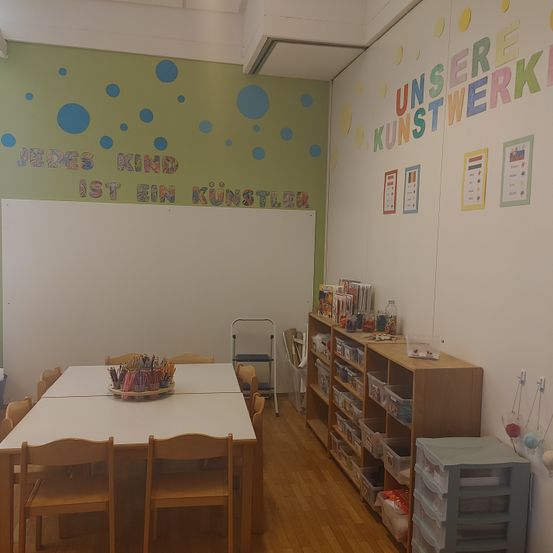 Ein Raum mit einem weißen Tisch und Stühlen, einer weißen Tafel an der Wand, einem Regal mit Büchern und anderen Gegenständen, einer Aufbewahrungsbox und einer farbenfrohen Wand mit Punkten und Text.