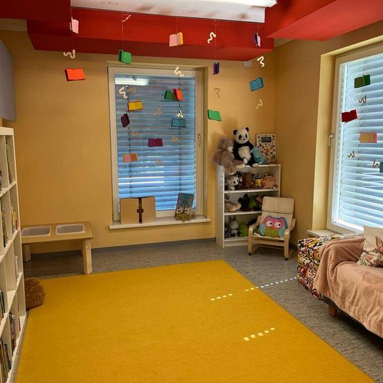 Ein gemütliches Zimmer mit einem gelben Teppich, einem Bücherregal, einem Schreibtisch, einem Fenster mit Jalousien, einem Stuhl, einem Regal mit Kuscheltieren und bunten Buchdekorationen, die von der Decke hängen.