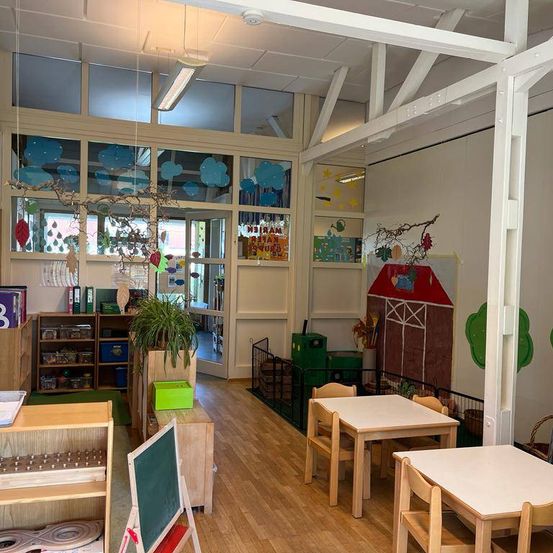 Ein Klassenzimmer mit Holzböden, weißen Wänden und Glasfenstern. Es hat Tische und Stühle, eine Topfpflanze und Regale mit Gegenständen. Eine Zeichnung eines Bauernhofs ist an der Wand, zusammen mit einer Tafel.