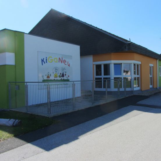 Ein modernes Gebäude mit grüner und oranger Wand, schwarzem Dach und Metallzaun davor. Das Gebäude ist mit dem Namen KiGaNeu beschriftet.