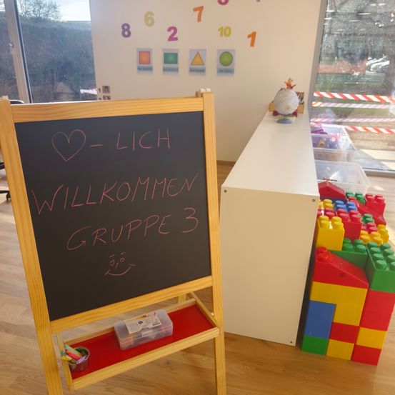 Ein Tafel mit der Botschaft 'Willkommen Gruppe 3' steht in einem Raum mit Holzböden. Ein Regal daneben hält einen Globus und Aufbewahrungsboxen. Bunte Bausteine sind in der Nähe.