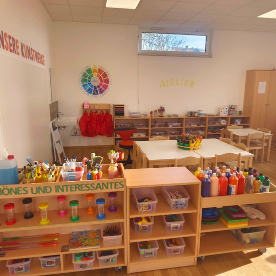 Ein Klassenzimmer mit Holzregalen, die verschiedene Bastelmaterialien enthalten, einem Tisch mit einem Farbkreis und einer Wand mit einem Gemälde von Bäumen und dem Wort Atelier.