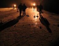 Mehrere Menschen stehen nachts auf einer vereisten Oberfläche und bereiten sich auf eine Partie Curling vor. Sie tragen Winterkleidung und halten Curlingbesen. Es gibt zwei gelbe Ziele auf dem Eis.