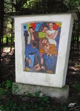 Ein Mosaikbild, das mehrere Menschen in verschiedenen Posen darstellt, ist in einem weißen Rahmen auf einem Betonblock in einem bewaldeten Gebiet montiert.
