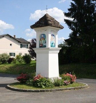 Eine weiße Struktur mit einem braunen Dach steht in einem Grasbereich, umgeben von Blumen und Pflanzen. Ein Haus mit Schornstein und Fenstern ist im Hintergrund, mit Bäumen und Büschen. Der Himmel ist blau mit weißen Wolken.