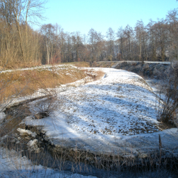 Eine verschneite Landschaft mit einem mäandernden Fluss, umgeben von laublosen Bäumen, unter einem klaren blauen Himmel.