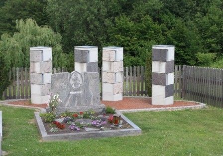 Ein Denkmal in einem Garten mit einer zentralen Plakette und vier Steinsäulen. Blumen und Pflanzen umgeben die Plakette, und ein Holzzaun begrenzt das Gebiet. Bäume sind im Hintergrund.