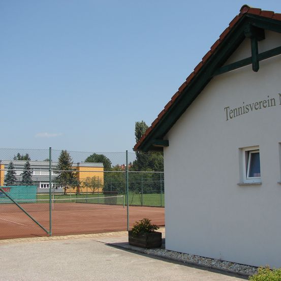 Tennisverein Gebäude mit einem Tennisplatz, der durch einen Zaun sichtbar ist, unter einem klaren blauen Himmel mit grünen Bäumen im Hintergrund.