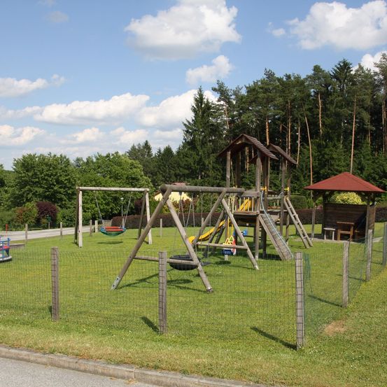 Ein Außenspielplatz mit Schaukeln, einer Rutsche und einem Reifenschaukel auf einem Rasenfeld, umgeben von einem Zaun und Bäumen.