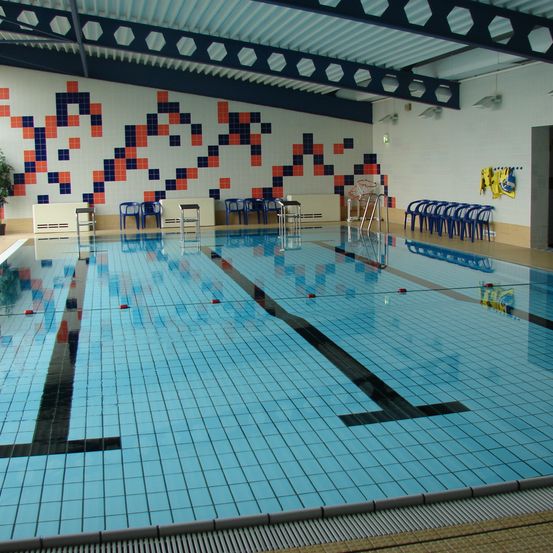 Ein Hallenschwimmbad mit blauen Fliesen und einem geometrischen Muster an den Wänden. Blaue Stühle sind auf beiden Seiten des Beckens angeordnet.
