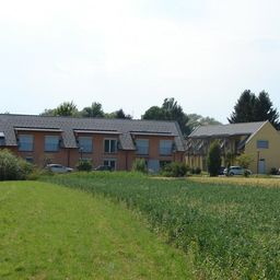 Ein Wohngebäude mit mehreren Fenstern und einem Solarpanel auf dem Dach, umgeben von einem großen Feld und mehreren geparkten Autos.