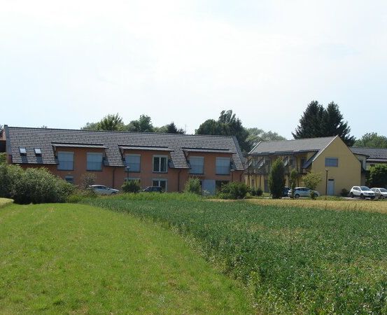 Ein Wohngebäude mit mehreren Fenstern und einem Solarpanel auf dem Dach, umgeben von einem großen Feld und mehreren geparkten Autos.