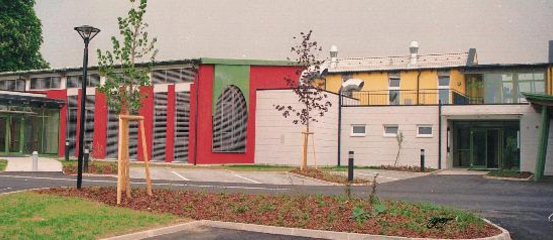Ein Außenansicht eines Gebäudes mit rot, gelb und grün gefärbten Wänden, mit Glasfenstern und Türen, mit Bäumen und einem geparkten Auto im Vordergrund.