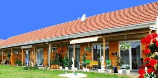 Ein Gebäude mit rotem Dach und weißen Wänden hat mehrere Fenster und eine weiße Markise. Die Vorderseite hat einen Garten mit roten Blumen und Topfpflanzen. In der Mitte steht eine Statue.