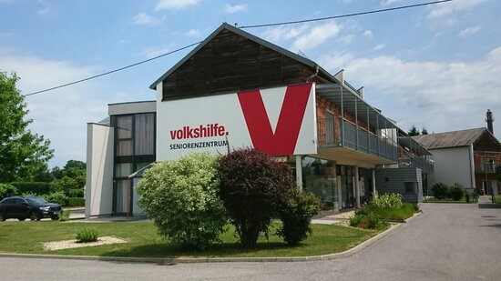 Ein Gebäude mit einem roten und weißen 'V'-Logo an der Seite, beschriftet mit 'volkshiffe. SENIORENZENTRUM'. Ein Auto ist davor geparkt, und das Gebäude ist von Grünpflanzen umgeben.