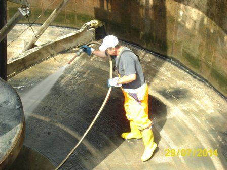 Ein Mann in Schutzkleidung reinigt mit einem Schlauch das Innere eines Betontanks. Das Datum 29/07/2014 ist in der unteren rechten Ecke sichtbar.