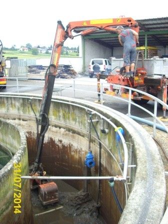 Ein Mann benutzt einen Kran, um Schlamm aus einem großen, rostigen Tank zu entnehmen. Der Kran steht auf einem Lastwagen und ist mit einer Metallkonstruktion mit Rohren verbunden. Ein weißer Van ist im Hintergrund geparkt.