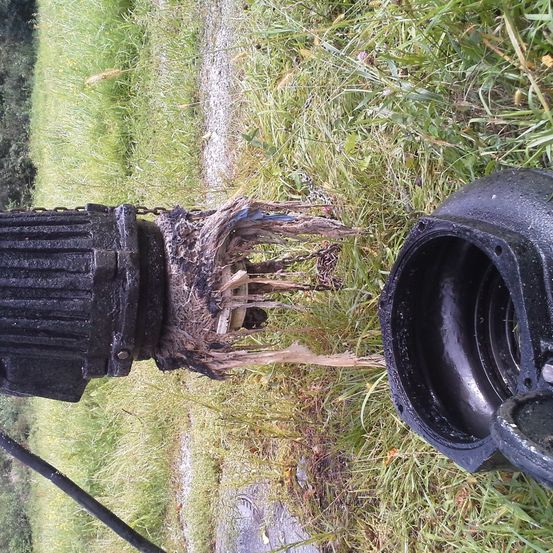 Eine schwarze Pumpe mit einer Kette ist kaputt und liegt auf dem Gras. Der Deckel ist abgenommen und zeigt das Innere.