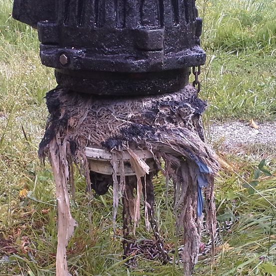 Ein verwittertes und verrostetes schwarzes Ventil ist an einer Kette befestigt. Es ist mit trockenem Gras und Ablagerungen bedeckt. Das Ventil steht im Freien auf einer Rasenfläche.