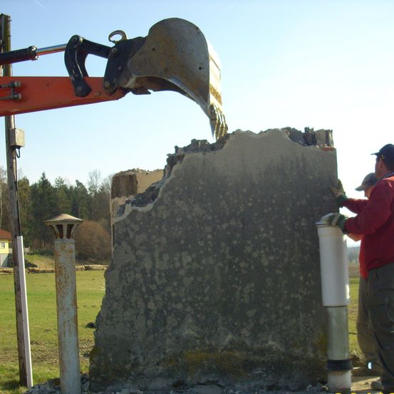 Zwei Männer arbeiten an einer großen Betonmauer. Einer hält einen weißen Behälter, und der andere berührt die Mauer. In der Nähe steht ein großer oranger Bagger mit einem großen Eimer. Hinter der Mauer befindet sich ein Feld und Bäume.
