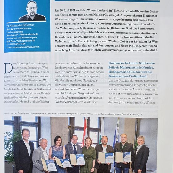 Das Bild zeigt eine Zertifikatsverleihungszeremonie mit mehreren Personen in formeller Kleidung, die Urkunden halten. Der Hintergrund ist innen und zeigt eine Flagge und ein Fenster. Eine Person in Anzug sticht in der Mitte hervor.