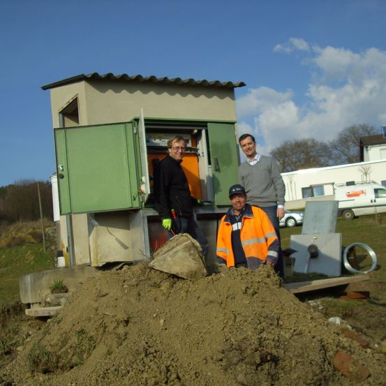 Drei Männer stehen an einem sonnigen Tag in der Nähe eines kleinen Gebäudes, möglicherweise eines Versorgungskastens. Der Mann in der Mitte hält eine Schaufel und lächelt.