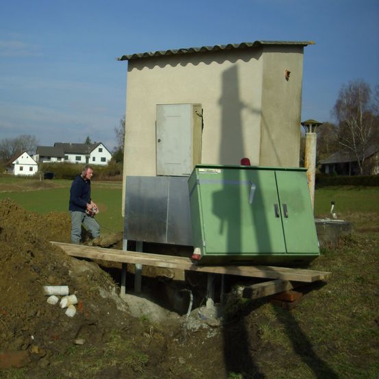 Ein Mann in einer blauen Jacke steht neben einem Gebäude mit einem grünen Schrank an der Seite. Das Gebäude befindet sich auf einem Grundstück.