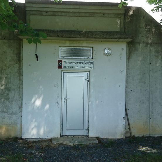 Ein Betongebäude mit einer weißen Tür und einem Schild, das Wasserversorgung Neudau Hochbehälter - Hackerberg besagt.