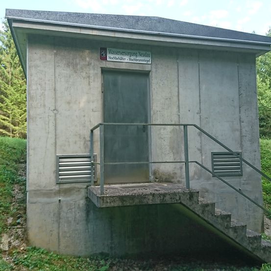 Ein Betongebäude mit einer Metalltür und einer Treppe. Ein Schild zeigt 'Wasserversorgung Neudau Hochbehälter Hochbrunnriegl' an.