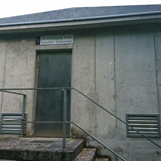 Ein Außenansicht eines Gebäudes mit einem Schild, das 'Wasserversorgung Neudau Hochbehälter - Hochbrunnriegl' über einer geschlossenen Tür und einem Metallgeländer auf den Stufen steht.