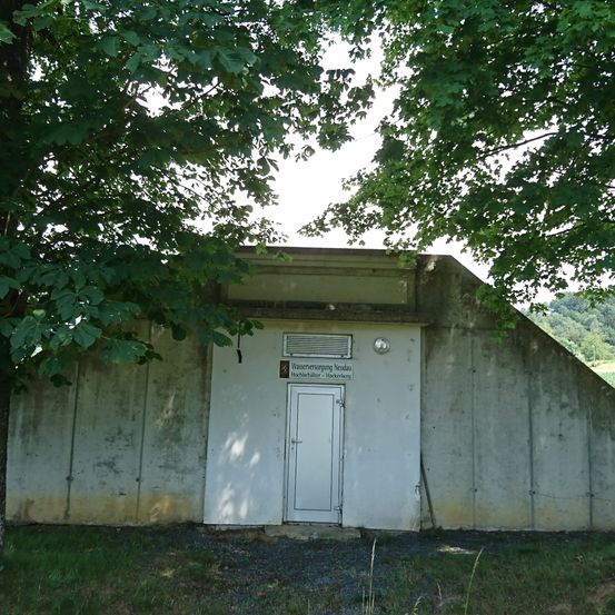 Ein altes Betongebäude mit einer weißen Tür und einem Schild mit der Aufschrift 'Wasserversorgung Neckau' befindet sich im Freien. Es gibt zwei Bäume in der Nähe.