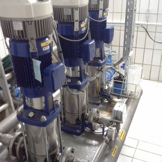 Mehrere industrielle blaue und silberne Pumpen sind auf einer metallischen Oberfläche montiert, verbunden mit Schläuchen und Rohren. In der Nähe befindet sich ein kleiner blauer Behälter mit Griff. Der Boden ist gefliest.