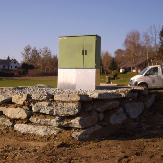 Ein grüner Stromkasten steht auf einem Betonsockel, umgeben von Steinen. Ein weißer Pickup-Truck ist in der Nähe geparkt. Im Hintergrund befinden sich Häuser und Bäume.