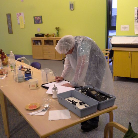 Eine Person in einer Plastikhülle schreibt auf einem Stück Papier auf einem Tisch. Es gibt verschiedene Gegenstände auf dem Tisch. Hinter der Person steht ein Holzschrank.