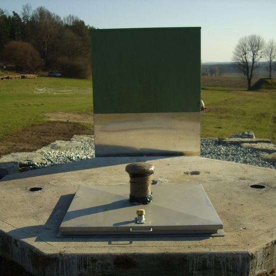 Ein grünes rechteckiges Objekt steht auf einer Betonplattform. Ein Metalldeckel ist auf der Plattform befestigt und mit einem Bolzen gesichert. Die Plattform ist von Kies umgeben, und ein Baum ist im Hintergrund zu sehen.