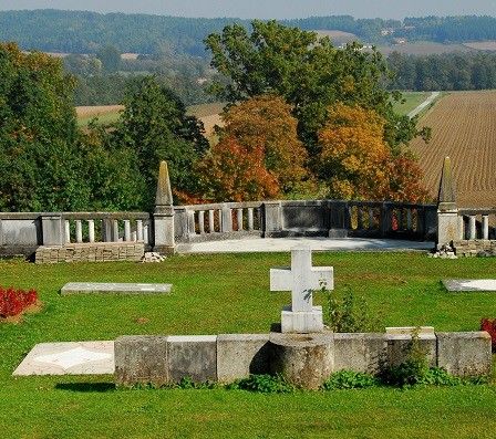 Ein ruhiger Friedhof mit Grabsteinen und Kreuzen, umgeben von einer Steinmauer, vor dem Hintergrund eines Feldes und Bäumen.