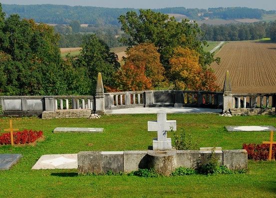 Ein ruhiger Friedhof mit Grabsteinen und Kreuzen, umgeben von einer Steinmauer, vor dem Hintergrund eines Feldes und Bäumen.