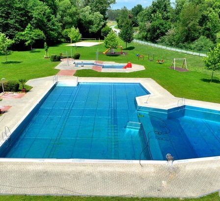 Eine Vogelperspektive eines großen Schwimmbeckens mit blauem Wasser, umgeben von grünem Gras, Bäumen und Büschen. Im Hintergrund befindet sich ein Spielplatz mit Rutsche und Schaukeln.