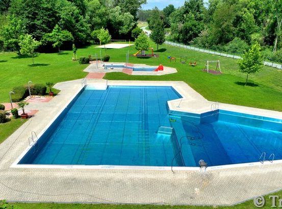 Eine Vogelperspektive eines großen Schwimmbeckens mit blauem Wasser, umgeben von grünem Gras, Bäumen und Büschen. Im Hintergrund befindet sich ein Spielplatz mit Rutsche und Schaukeln.