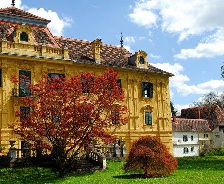Ein gelbes historisches Herrenhaus mit roten Herbstblättern davor, umgeben von grünem Gras und Bäumen, unter einem blauen Himmel mit Wolken.