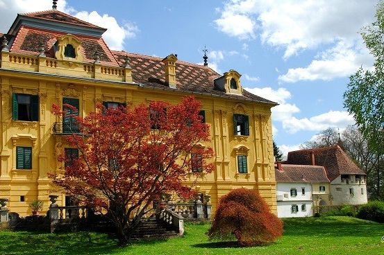 Ein gelbes historisches Herrenhaus mit roten Herbstblättern davor, umgeben von grünem Gras und Bäumen, unter einem blauen Himmel mit Wolken.